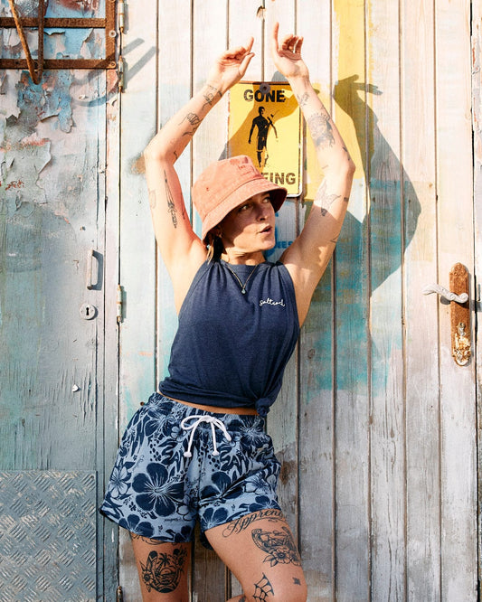 Person with tattoos wearing a Saltrock branded bucket hat, sleeveless shirt, and Soifra Hibiscus - Womens Sweat Short - Blue with a Hawaiian hibiscus design poses in front of a textured wooden wall with a "Gone Surfing" sign.