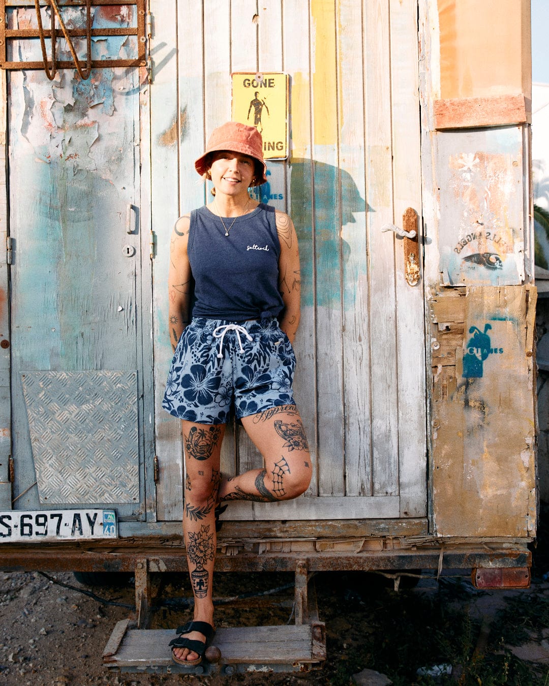 Person wearing a hat, sleeveless shirt, and Soifra Hibiscus - Womens Sweat Short - Blue by Saltrock, standing in front of a weathered wooden door with various posters and graffiti, leaning casually, and smiling.