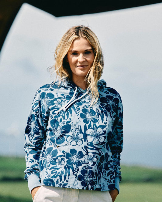 A person with long, wavy hair stands outside wearing the Saltrock Soifra Hibiscus - Womens Pop Hood in blue. The relaxed boxy cropped fit beautifully complements the Hawaiian hibiscus print on the hoodie. In the background, greenery and a cloudy sky create a serene atmosphere.