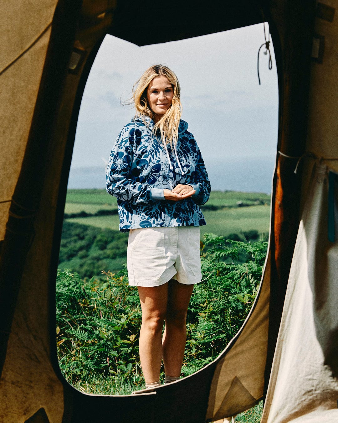 A person wearing a Saltrock Soifra Hibiscus - Women's Pop Hood in blue, featuring a relaxed boxy cropped fit with a Hawaiian hibiscus print, and white shorts stands in front of a green landscape, as seen through the frame of a tent entrance.