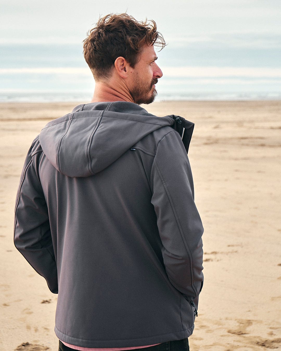 The back of a man standing on the beach wearing a Saltrock Munros - Mens Softshell Jacket - Grey, featuring water-resistant fabric and zip pockets.