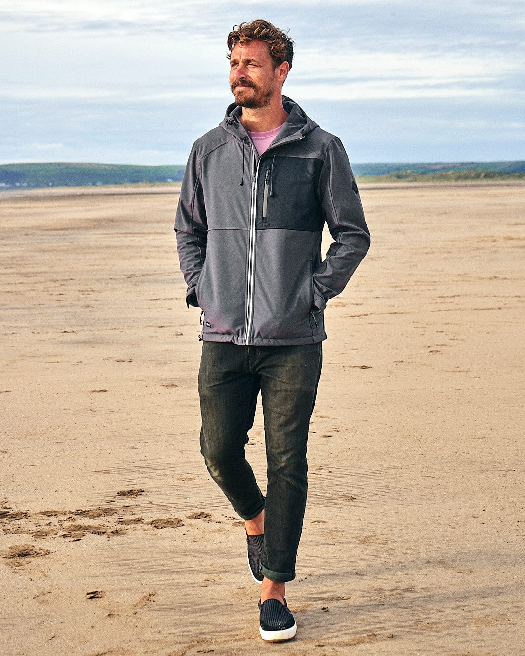 A man strolling along the beach in a Saltrock Munros - Mens Softshell Jacket - Grey, which features zip pockets and is made of water-resistant fabric.
