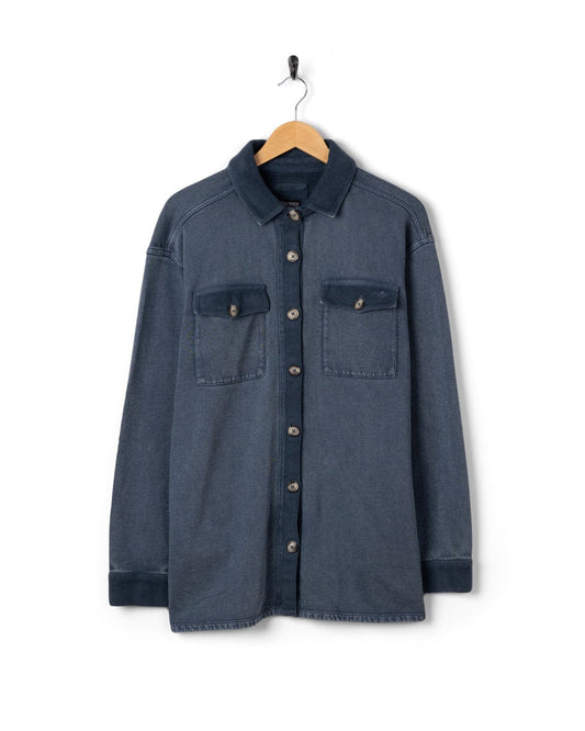 The Maddie Womens Oversized Shirt by Saltrock features long sleeves, two chest pockets, and a blue denim finish. Hanging on a wooden hanger against a white background, it's ideal as an acid wash over-shirt for effortless style.
