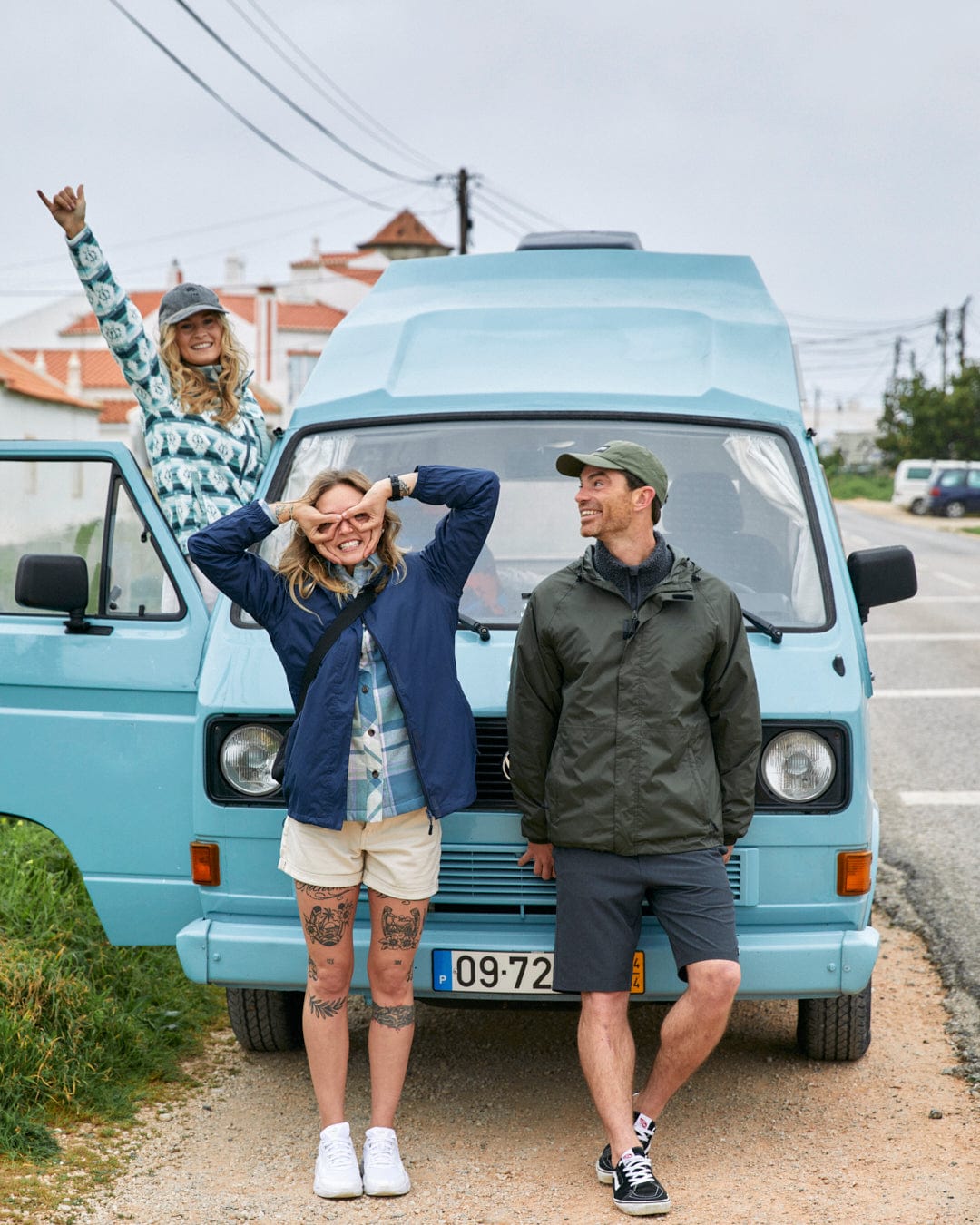 Three friends posing joyfully in front of a light blue vintage van on a cloudy day; two women and one man, with the man wearing a Saltrock Rainier - Mens Packable Waterproof Jacket in Green.