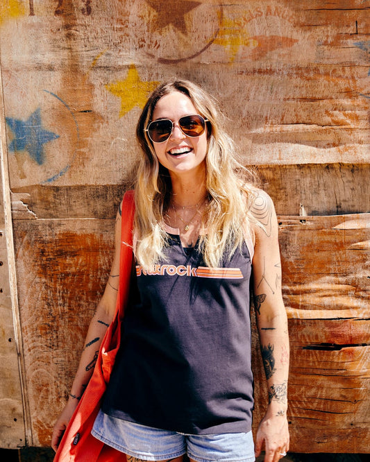 A smiling woman with sunglasses, wearing a Saltrock Retro Ribbon - Womens Vest in Dark Grey and a red cover-up made of 100% cotton, stands in front of a wooden wall with a painted star.