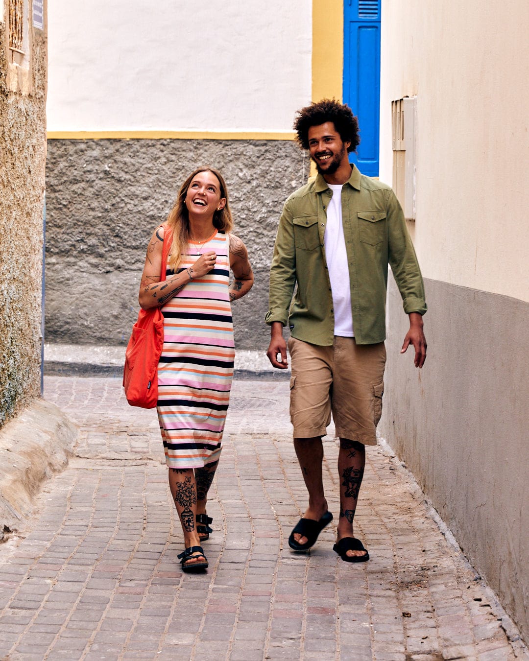 A man and a woman walking and laughing together down a narrow alley, the woman holding a drink and wearing a Saltrock Penare - Mens Long Sleeve Shirt - Green.