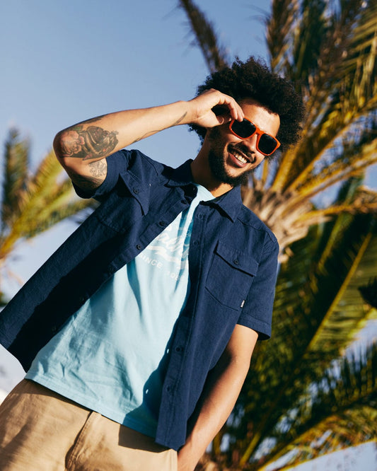 A stylish young man with an afro, wearing sunglasses and a Saltrock Polperro Men's Short Sleeve Shirt in Blue, stands outdoors with palm trees in the background.