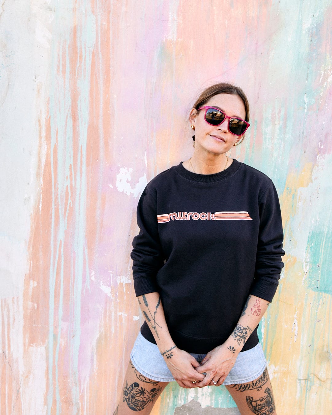 A woman in a Saltrock Retro Ribbon Tape - Womens Sweatshirt - Dark Grey and sunglasses stands confidently against a colorful, abstract mural.