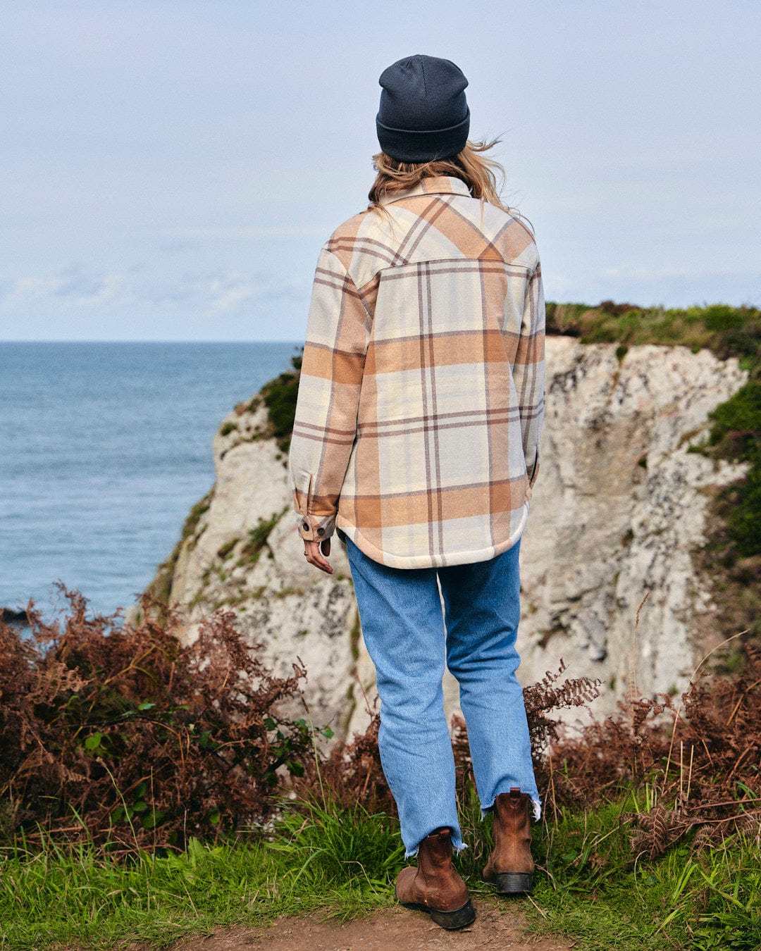 A man standing on a cliff overlooking the ocean wearing a Saltrock Stella - Womens Checked Borg Lined Shacket - Cream with buttoned cuffs plaid shirt.