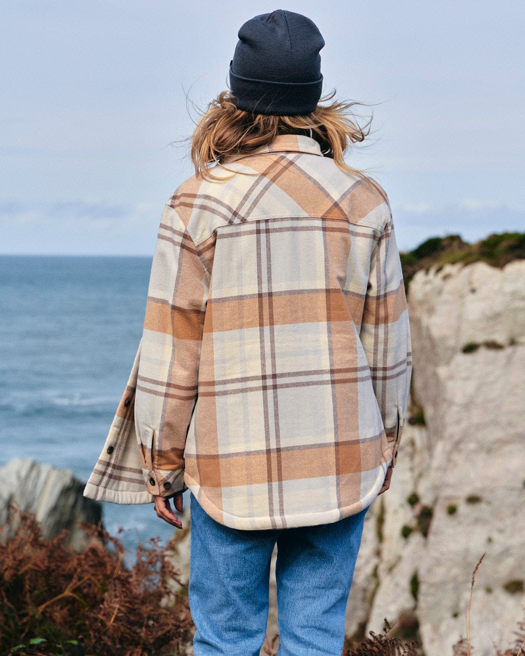 A woman wearing a Saltrock Stella - Womens Checked Borg Lined Shacket - Cream with buttoned cuffs, standing on a cliff overlooking the ocean.
