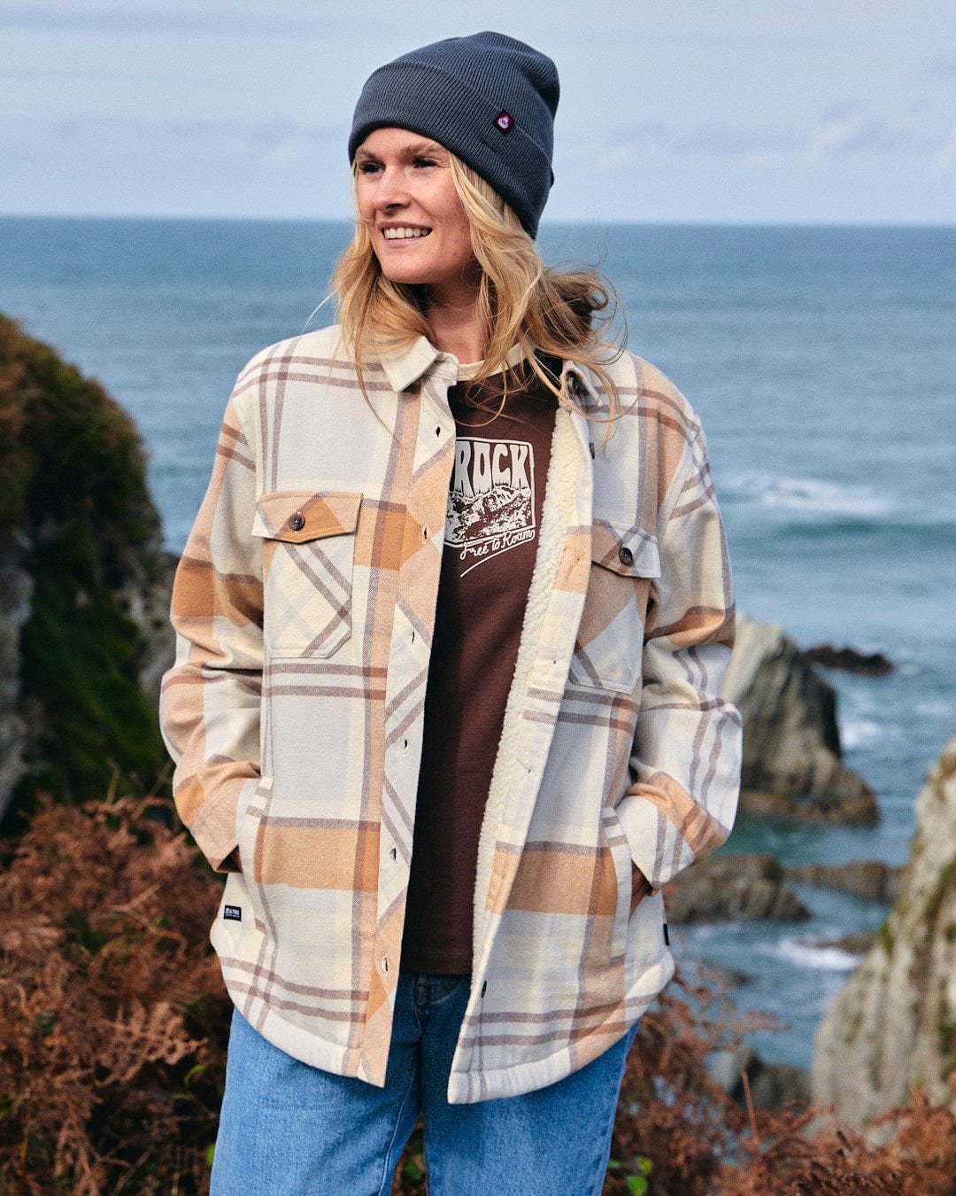 A woman wearing the Saltrock Stella - Womens Checked Borg Lined Shacket in Cream and beanie standing by the ocean.