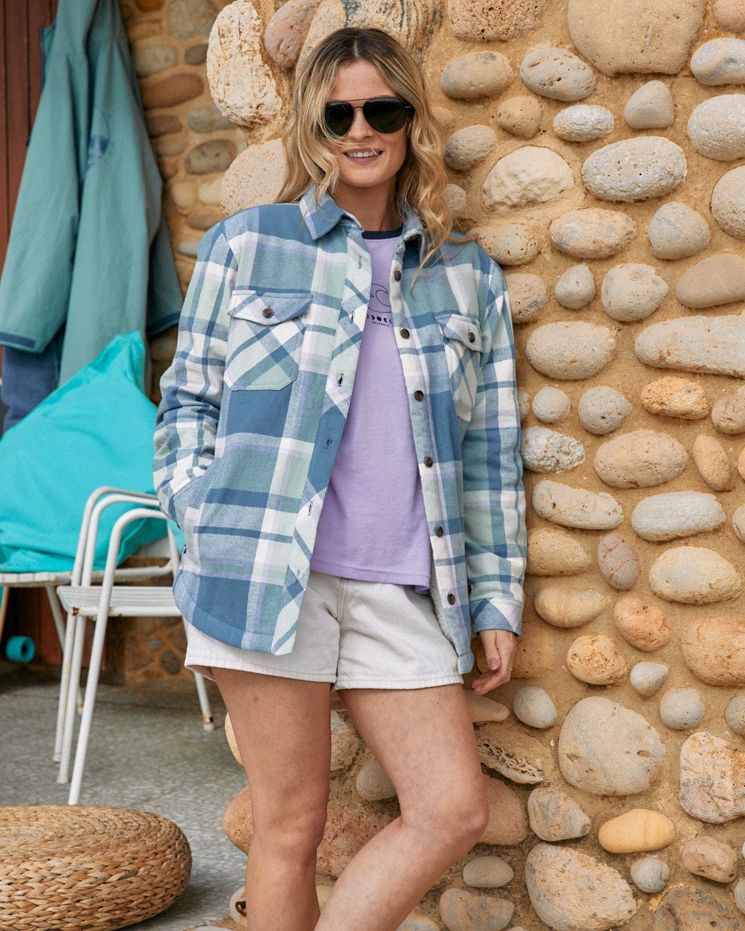 A woman wearing sunglasses stands against a cobblestone wall, donned in a casual Stella - Womens Borg Lined Long Sleeve Shirt in Blue over a purple t-shirt, paired with white shorts.