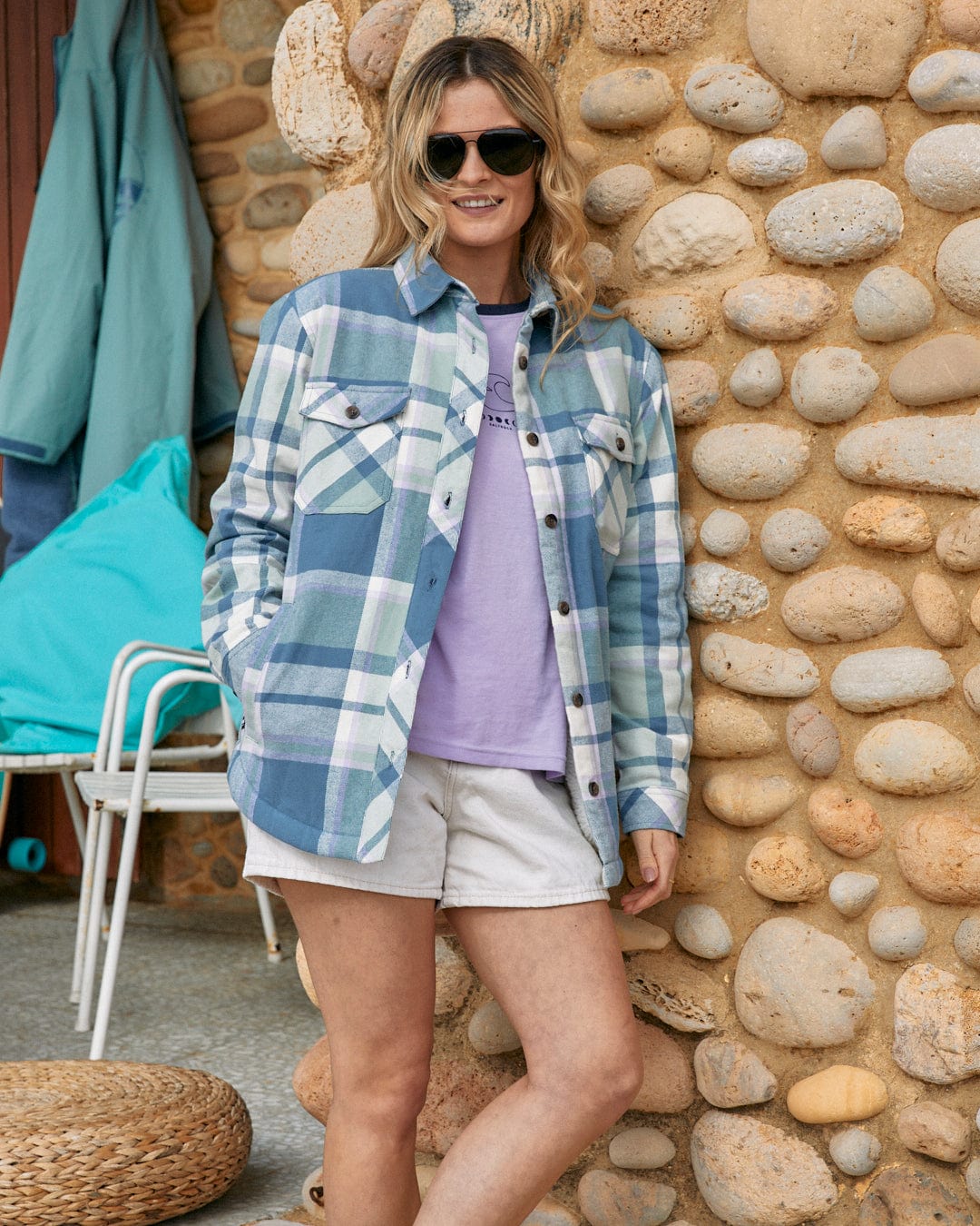 Woman in sunglasses and layered outfit with plaid jacket, Stella - Womens Borg Lined Long Sleeve Shirt in Blue by Saltrock, button-down shirt, and white shorts standing against a textured stone wall.