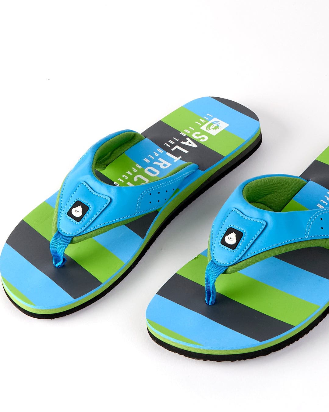 Saltrock's Striped Men's Flip Flops in blue and green feature an ergonomic upper, a cartoon face on the straps, and a grippy outsole, all set against a crisp white background.