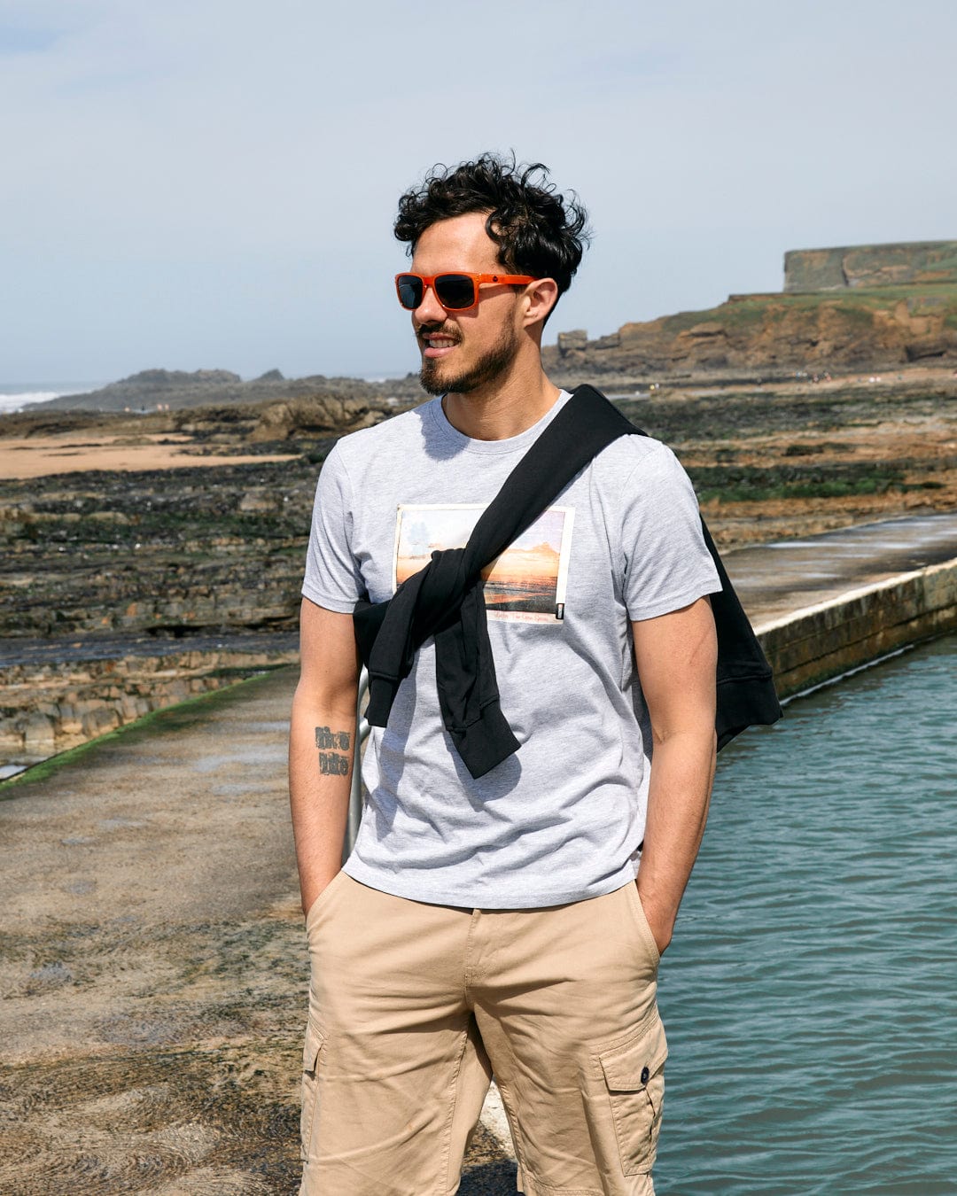 A man in sunglasses, wearing a Saltrock "Sun Sets" mens short sleeve t-shirt in grey marl with a crew neckline and khaki shorts, stands by a seaside with a rocky backdrop, smiling slightly.