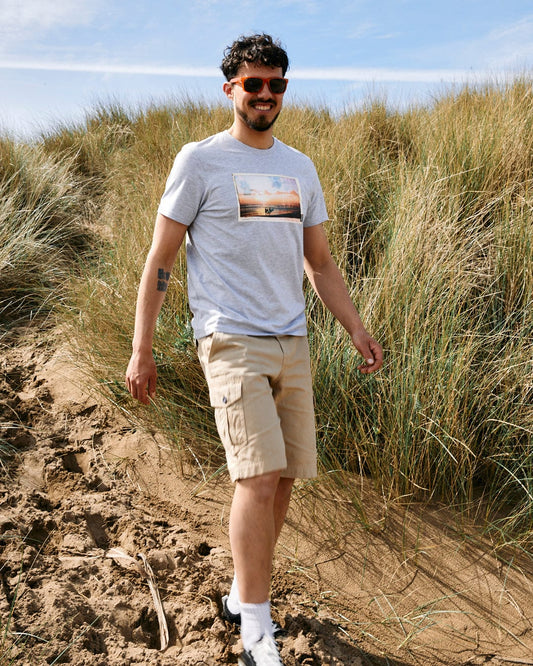 Man in sunglasses, Sun Sets - Mens Short Sleeve T-Shirt - Grey Marl by Saltrock, and khaki shorts walking on a sandy path through tall grass.