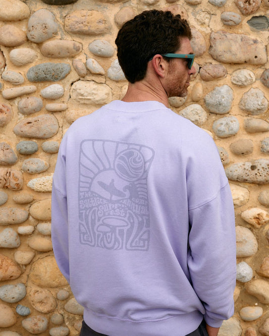 A man in sunglasses and a Saltrock Surf Fest - Mens Oversized Sweatshirt in Lilac stands in front of a stone wall.