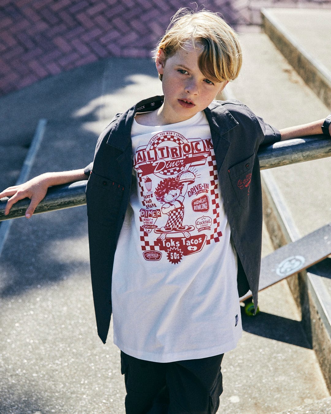 A young boy with blonde hair stands outside, leaning on a railing. He is wearing a Kingpin Krew - Kids Short Sleeve Shirt - Dark Grey by Saltrock and a black jacket draped over his shoulders, reminiscent of the rugged utility worker style.