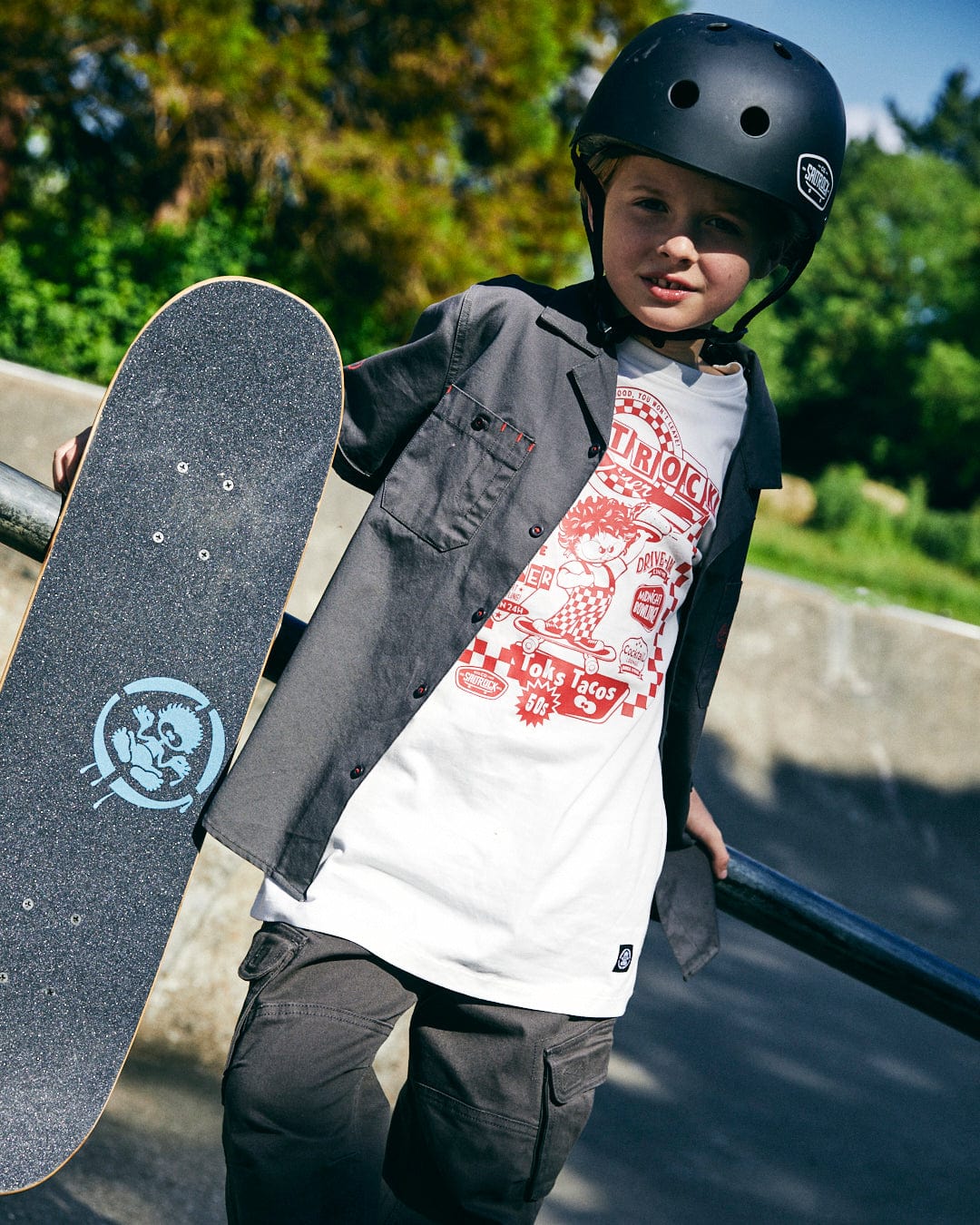 A young boy wearing a helmet and casual clothing with a Saltrock Kingpin Krew - Kids Short Sleeve Shirt - Dark Grey stands outdoors, holding his skateboard.