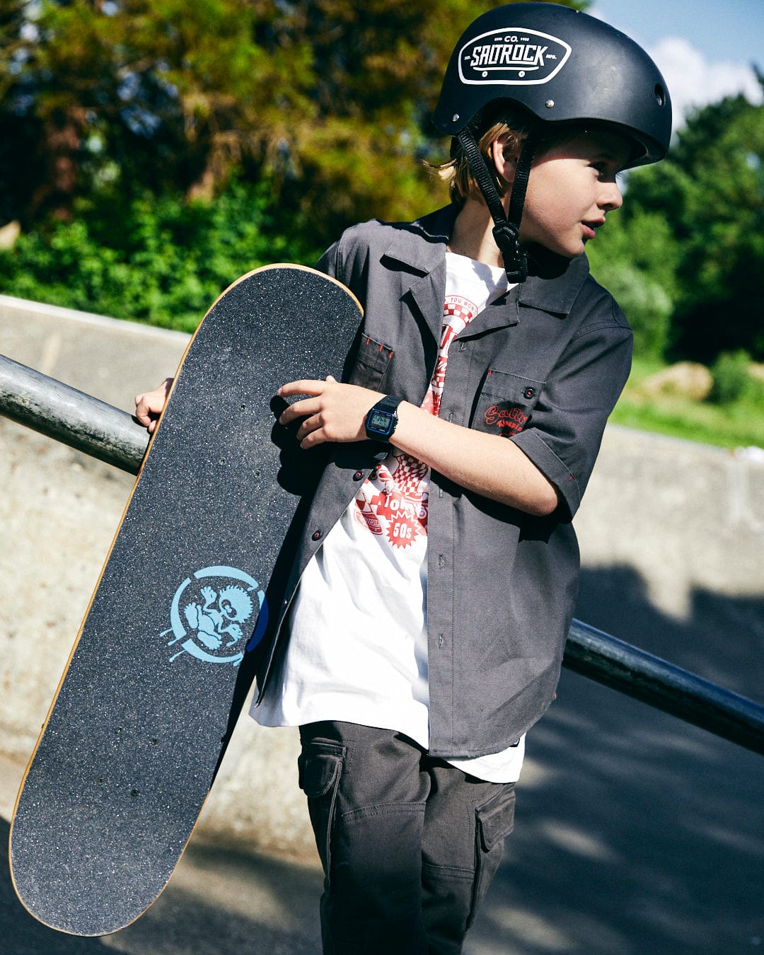 A young boy wearing a helmet and a Saltrock Kingpin Krew - Kids Short Sleeve Shirt - Dark Grey holds his skateboard while standing next to a metal railing at a skate park.
