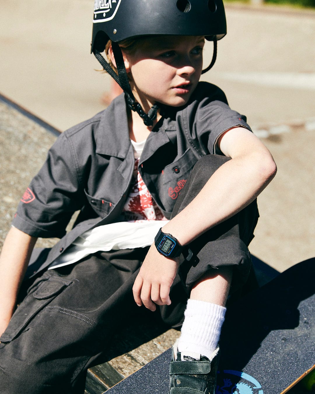 A young person wearing a black helmet and a Saltrock Kingpin Krew - Kids Short Sleeve Shirt - Dark Grey sits on a skateboard ramp, resting their hand on a skateboard featuring the Saltrock skateboard logo. They are looking to the side and wearing a digital wristwatch.