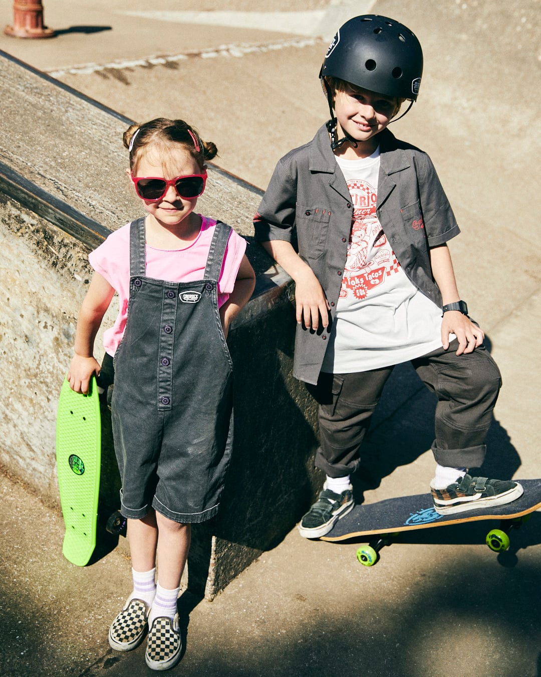 Two young children at a skatepark. One wears gray overalls in a utility worker style, pink sunglasses, and holds a green skateboard with the Saltrock skateboard logo. The other wears a black helmet, Kingpin Krew - Kids Short Sleeve Shirt - Dark Grey from Saltrock, black pants, and stands with their skateboard.