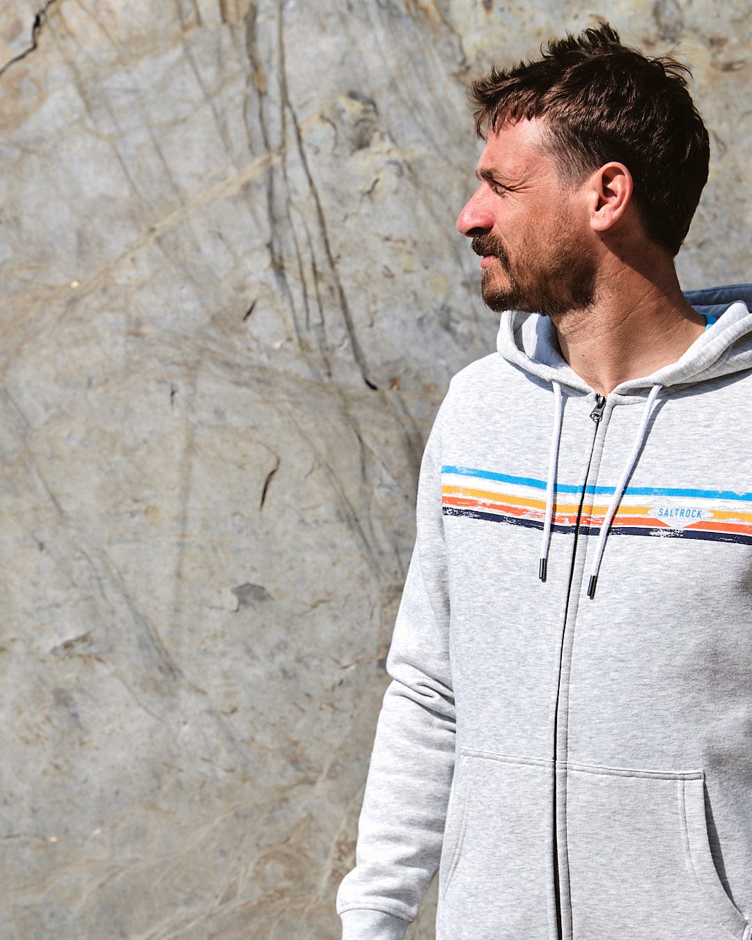 A man wearing a Saltrock Taped Stripe - Mens Zip Hoodie - Grey standing in front of a rock.