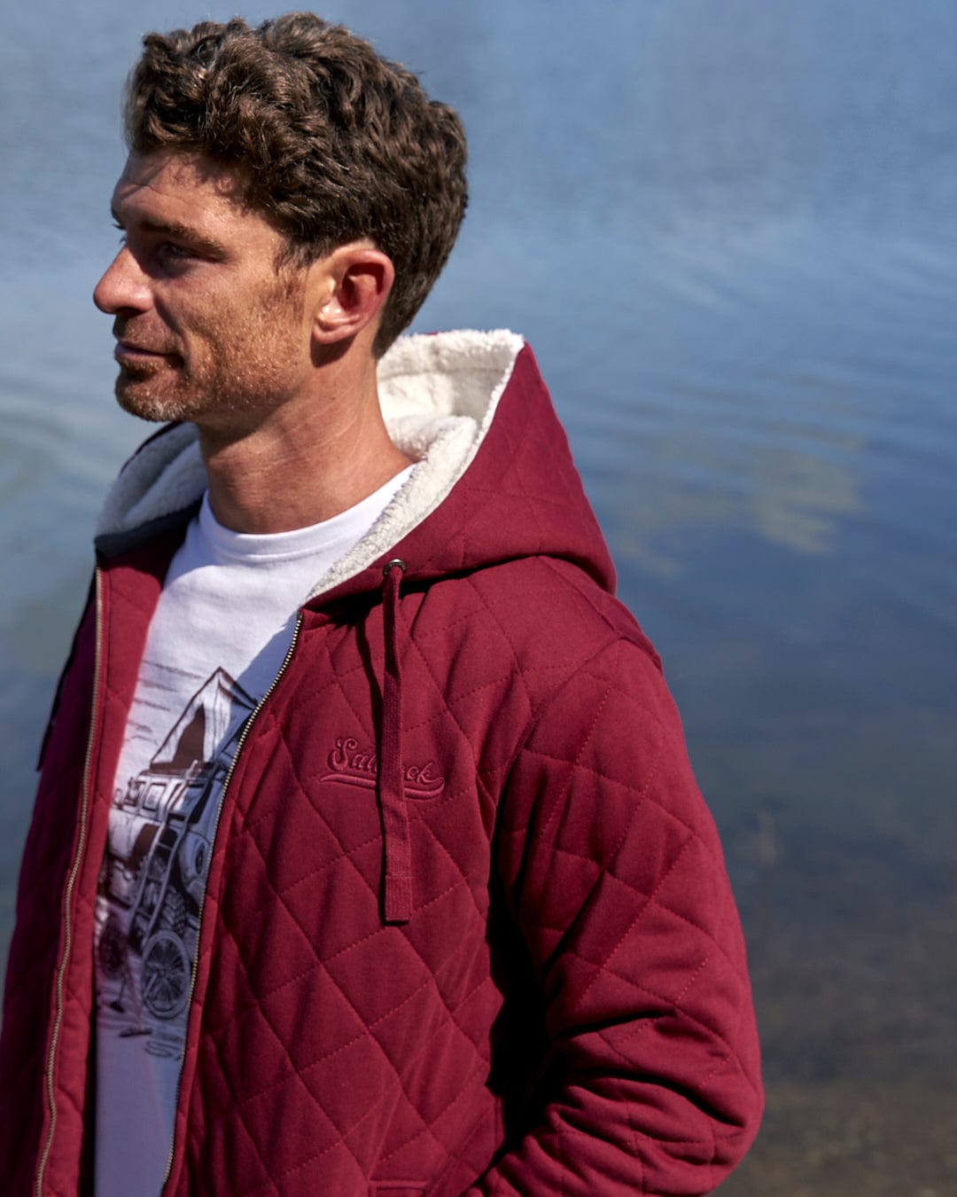 A man wearing a burgundy Saltrock Tarka - Mens Quilted Borg Lined Hoody - Red, a quilted fabric jacket, standing by a body of water.