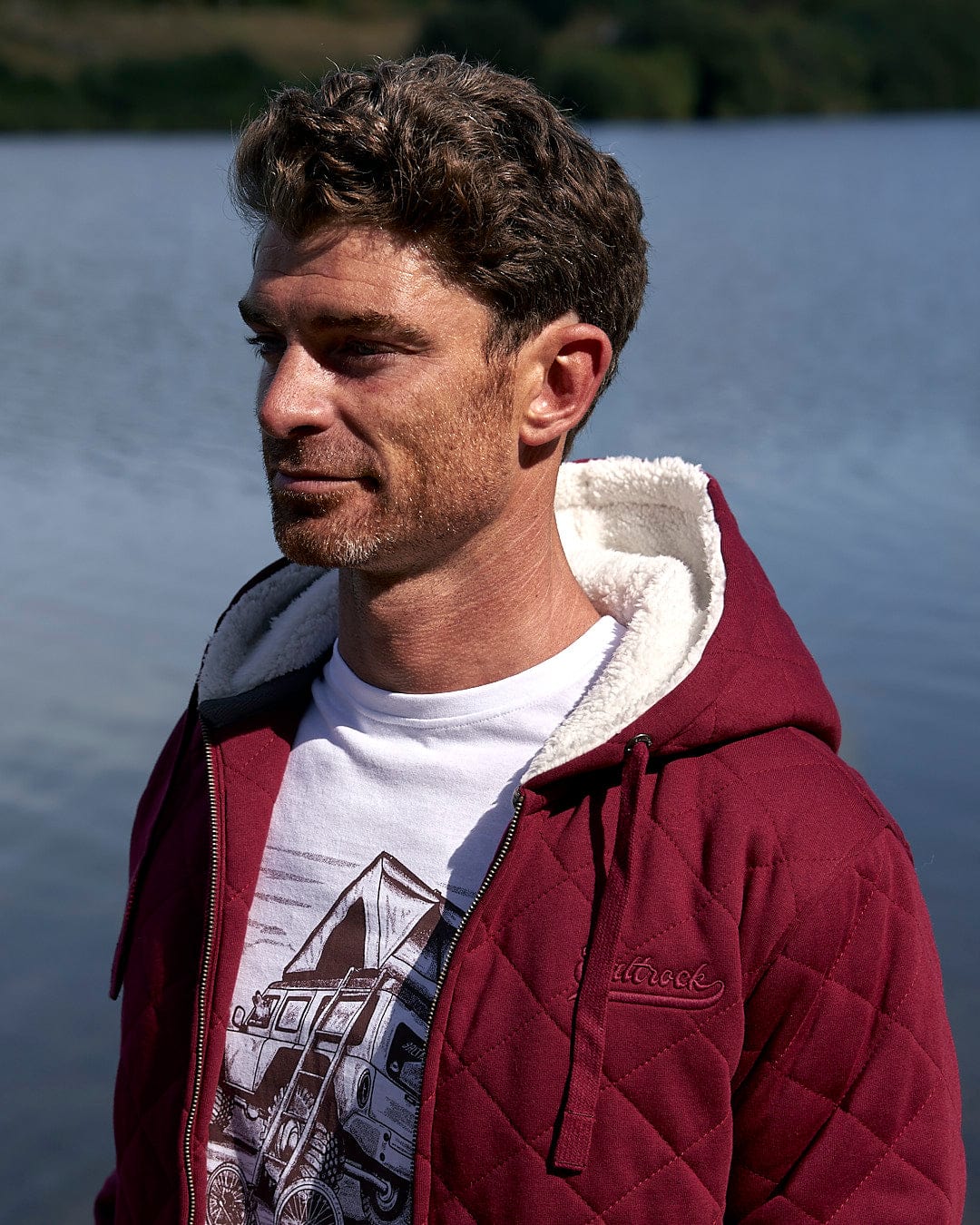 A man wearing a burgundy quilted jacket, showcasing the Saltrock branding, standing by a lake.