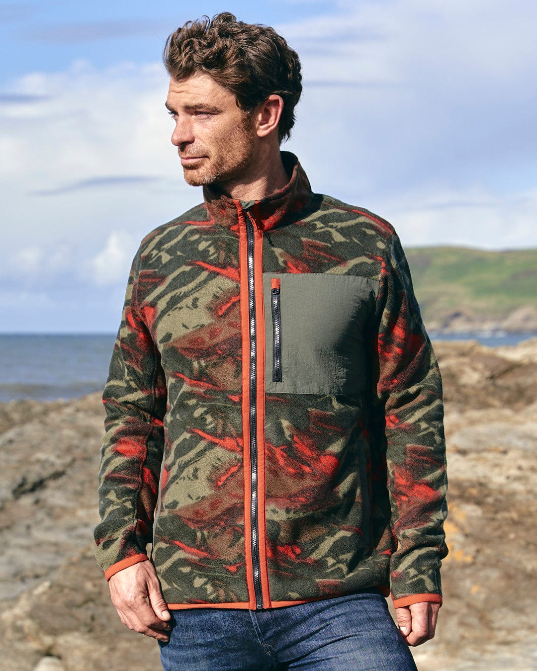 A man wearing a Terran - Mens Zip Thru Fleece - Green/Red jacket with a Saltrock badge on the beach.