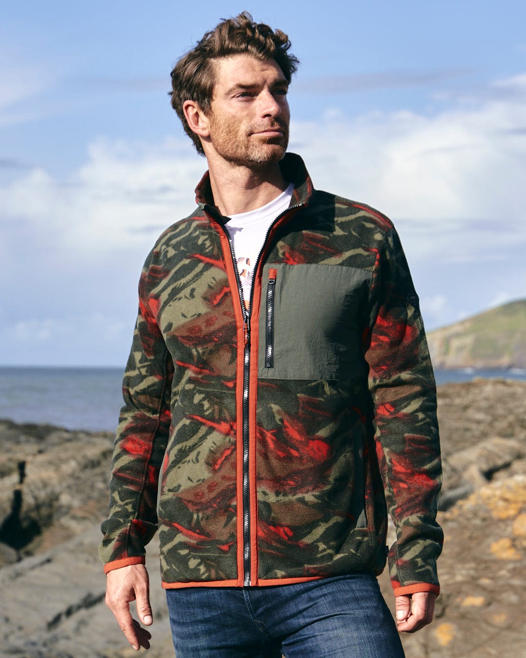 A man wearing a Saltrock Terran - Mens Zip Thru Fleece - Green/Red jacket (camo print) standing on a rocky beach.