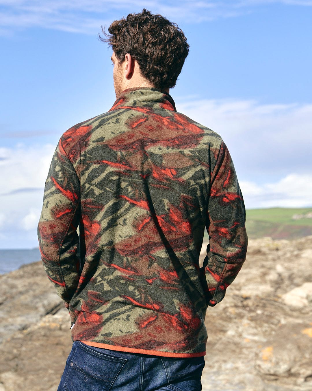 A man wearing a Saltrock Terran - Mens Zip Thru Fleece - Green/Red jacket with a utility chest pocket, looking at the ocean.