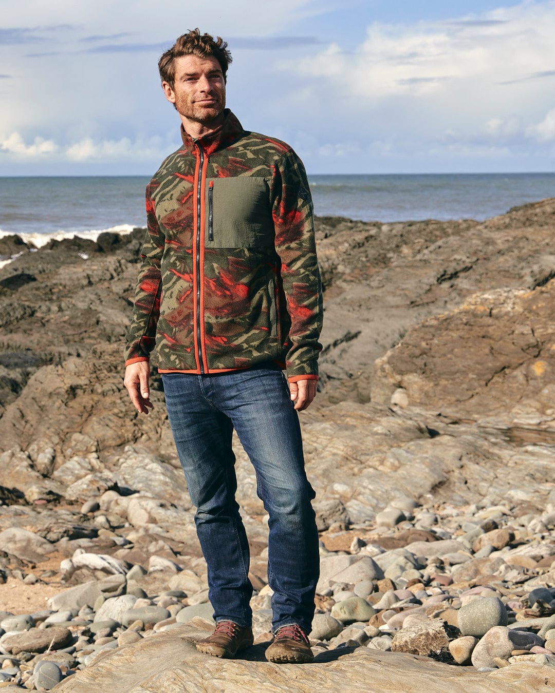 A man wearing camo print stands on a rocky beach, proudly showcasing his Saltrock Terran - Mens Zip Thru Fleece - Green/Red badge.