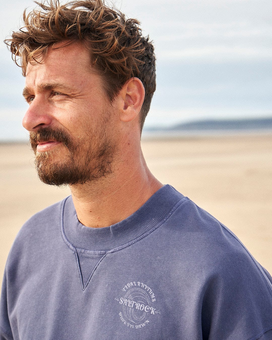 A man wearing a Tidal Rhythms - Mens Oversized Crew Sweat - Dark Blue sweatshirt by Saltrock standing on the beach.