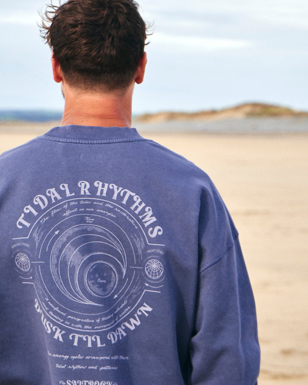 A man wearing an oversized dark blue Saltrock Tidal Rhythms - Mens Oversized Crew Sweatshirt stands on the beach, capturing the essence of Tidal Rhythms.