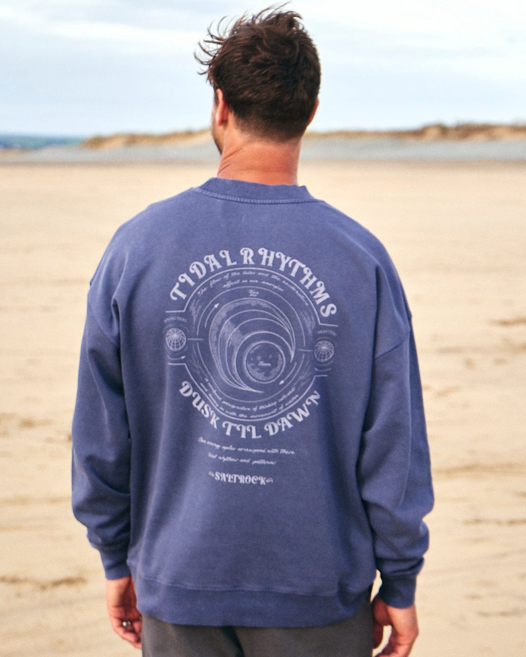 A man is standing on the beach wearing a Saltrock Tidal Rhythms - Mens Oversized Crew Sweat - Dark Blue.