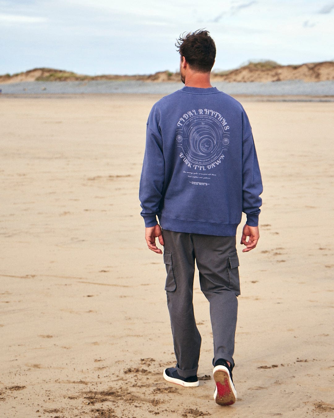 A man walking on the beach wearing a Saltrock Tidal Rhythms - Mens Oversized Crew Sweat - Dark Blue.