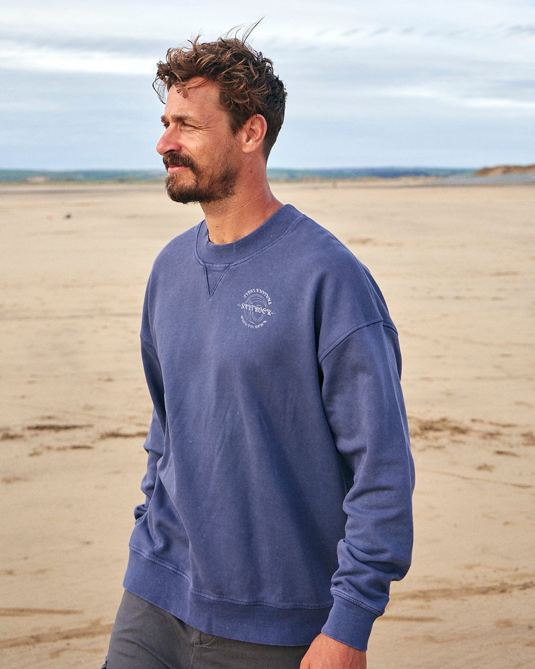 A man wearing a Tidal Rhythms - Mens Oversized Crew Sweat - Dark Blue by Saltrock, standing on the beach.