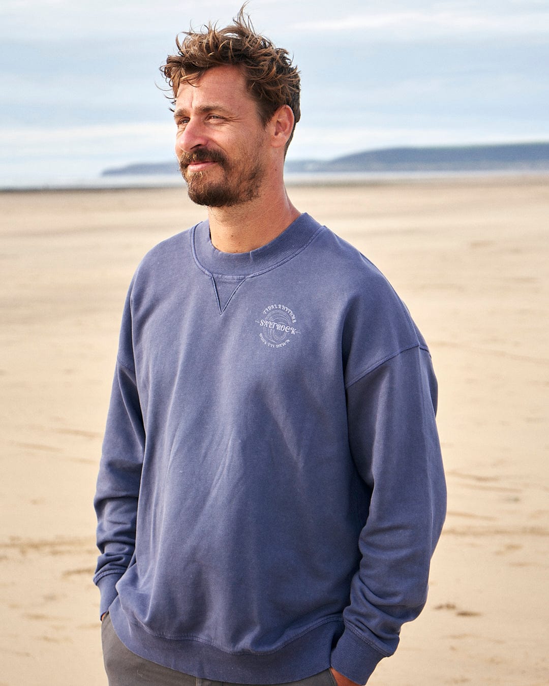 A man wearing the Saltrock Tidal Rhythms - Mens Oversized Crew Sweat in Dark Blue standing on the beach.
