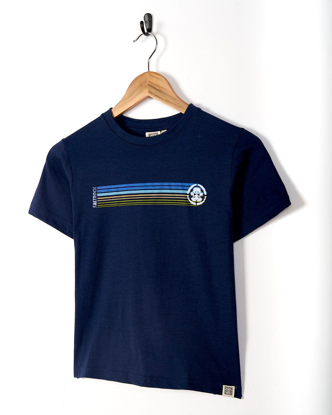 A Tok Stripe - Kids Short Sleeve T-Shirt in Dark Blue with retro stripes and a circular logo on the chest, hanging on a wall-mounted hook by Saltrock.