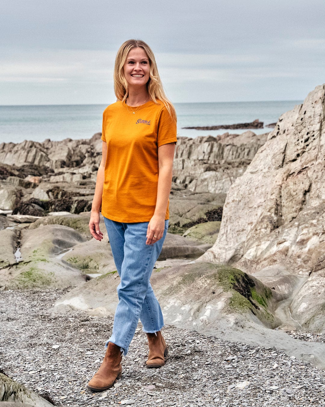 A woman in a Saltrock Womens Short Sleeve T-Shirt - Yellow, made of lightweight fabric, standing on a rocky beach.