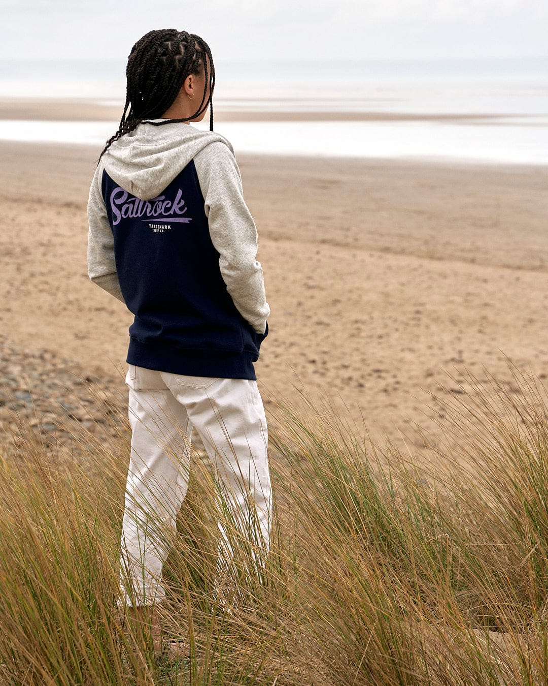 A girl is standing on a beach looking at the ocean wearing a Saltrock Trademark - Zip Hoodie in Dark Blue.