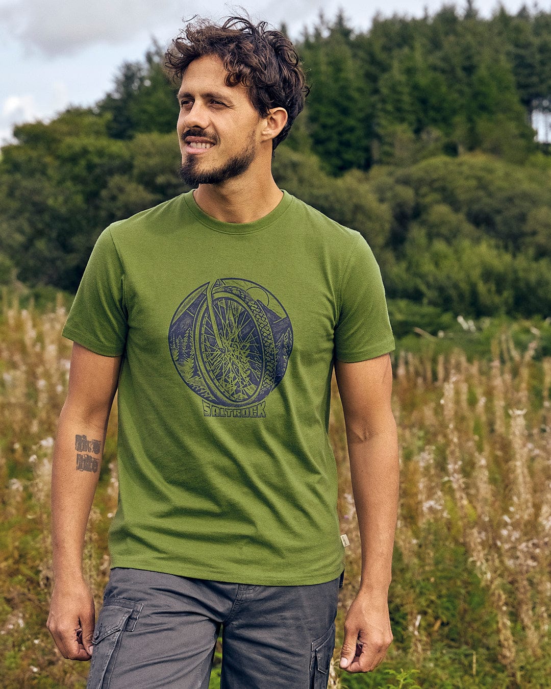 A man wearing a Saltrock Trail Bike - Mens Short Sleeve T-Shirt - Green stands in a field.