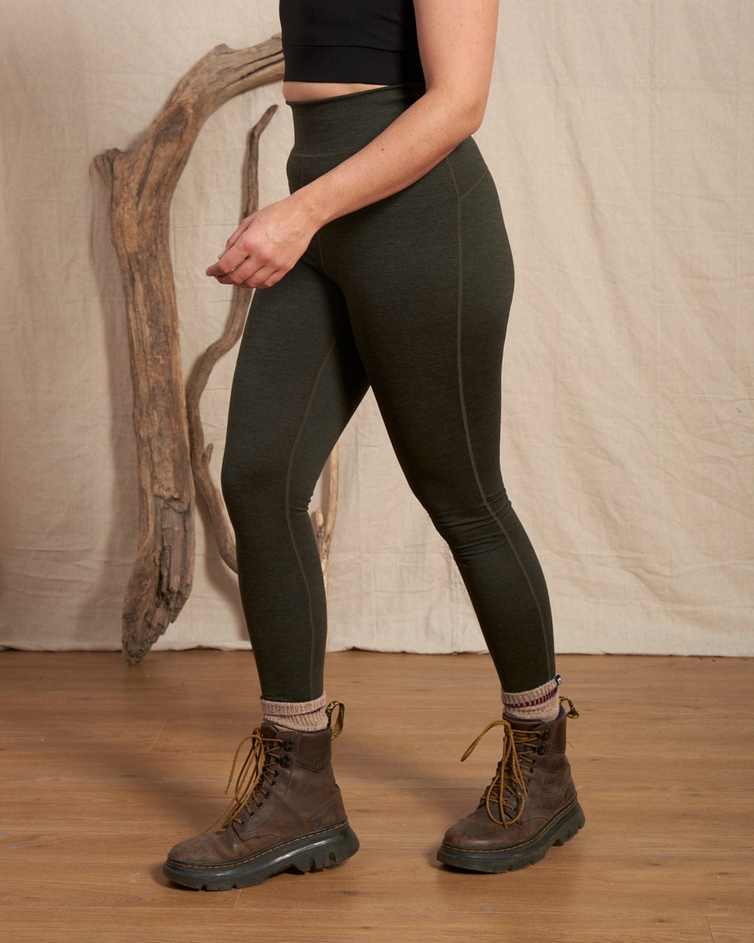 A person wears Saltrock Trek Women's 4 Way Stretch Outdoor Leggings in dark green, featuring a side thigh zipped pocket, with a black crop top, thick socks, and brown lace-up boots on a wooden floor with driftwood and a light backdrop.