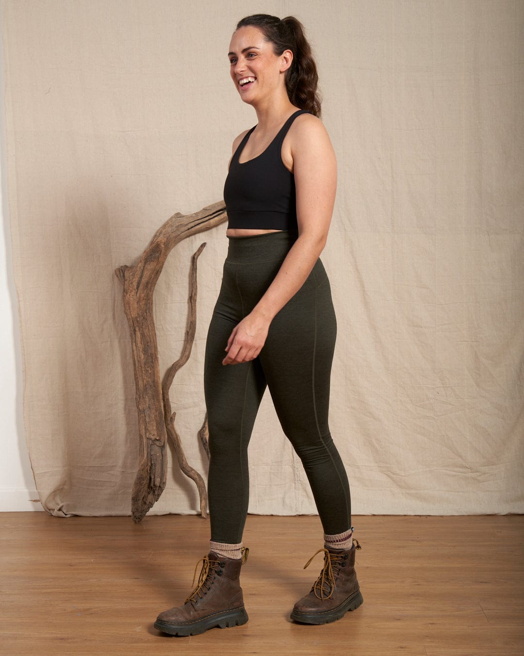 A woman wears a black crop top, Saltrock Trek Womens 4 Way Stretch Outdoor Leggings in Dark Green with a side thigh zipped pocket, and brown boots, standing indoors on a wooden floor before a beige backdrop and driftwood.