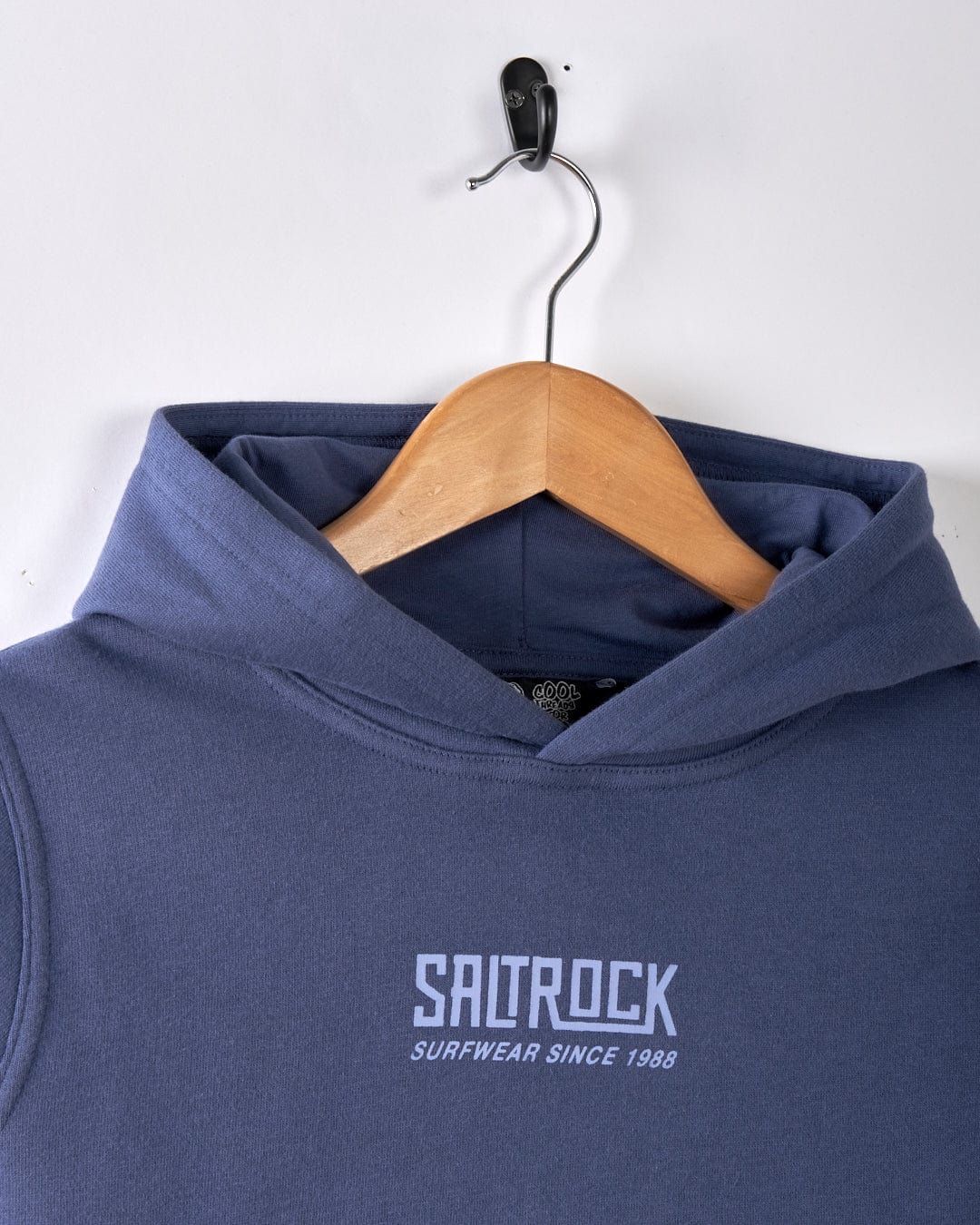 The Saltrock Tribal SR Kids Recycled Pop Hoodie in blue features "SURFWEAR SINCE 1988" text and a cozy kangaroo pocket. It hangs on a wooden hanger against a white background, highlighting its style and sustainable use of recycled materials.