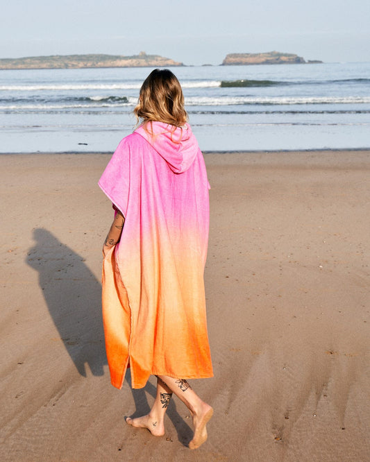 A woman draped in a Saltrock Tropic Dip - Changing Towel - Pink/Orange walks on a sandy beach, facing the ocean and distant cliffs.
