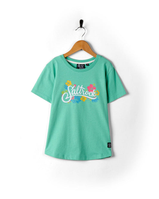 A green Tropic - Recycled Kids Short Sleeve T-Shirt with colorful "Saltrock print" logo, hanging on a wall-mounted hook against a white background.