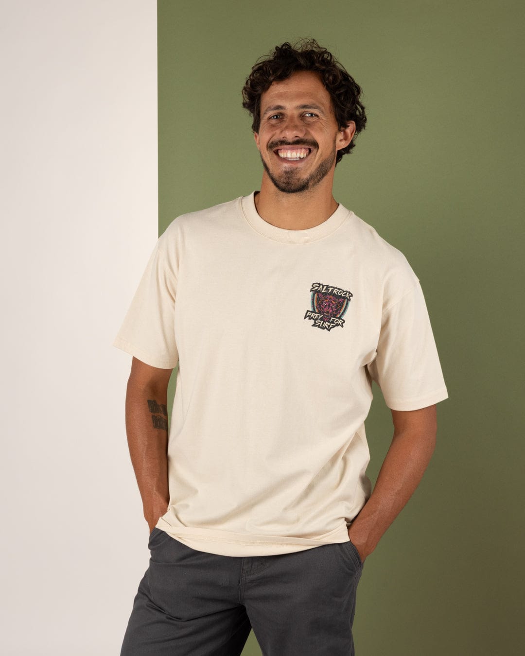 A man with curly hair and a beard smiles, standing against a split white and green background, wearing the Saltrock Wolfscale II Men's Oversized Short Sleeve T-Shirt in cream and dark pants with his hands in his pockets.