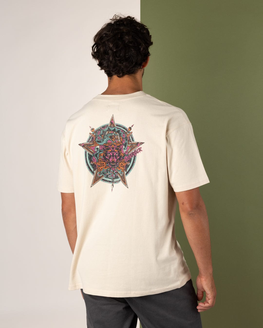 A man faces away, wearing the Saltrock Wolfscale II Men's Oversized Cream T-Shirt, made from 100% cotton and featuring a vibrant star and dragon design on the back. The split background is green and off-white.